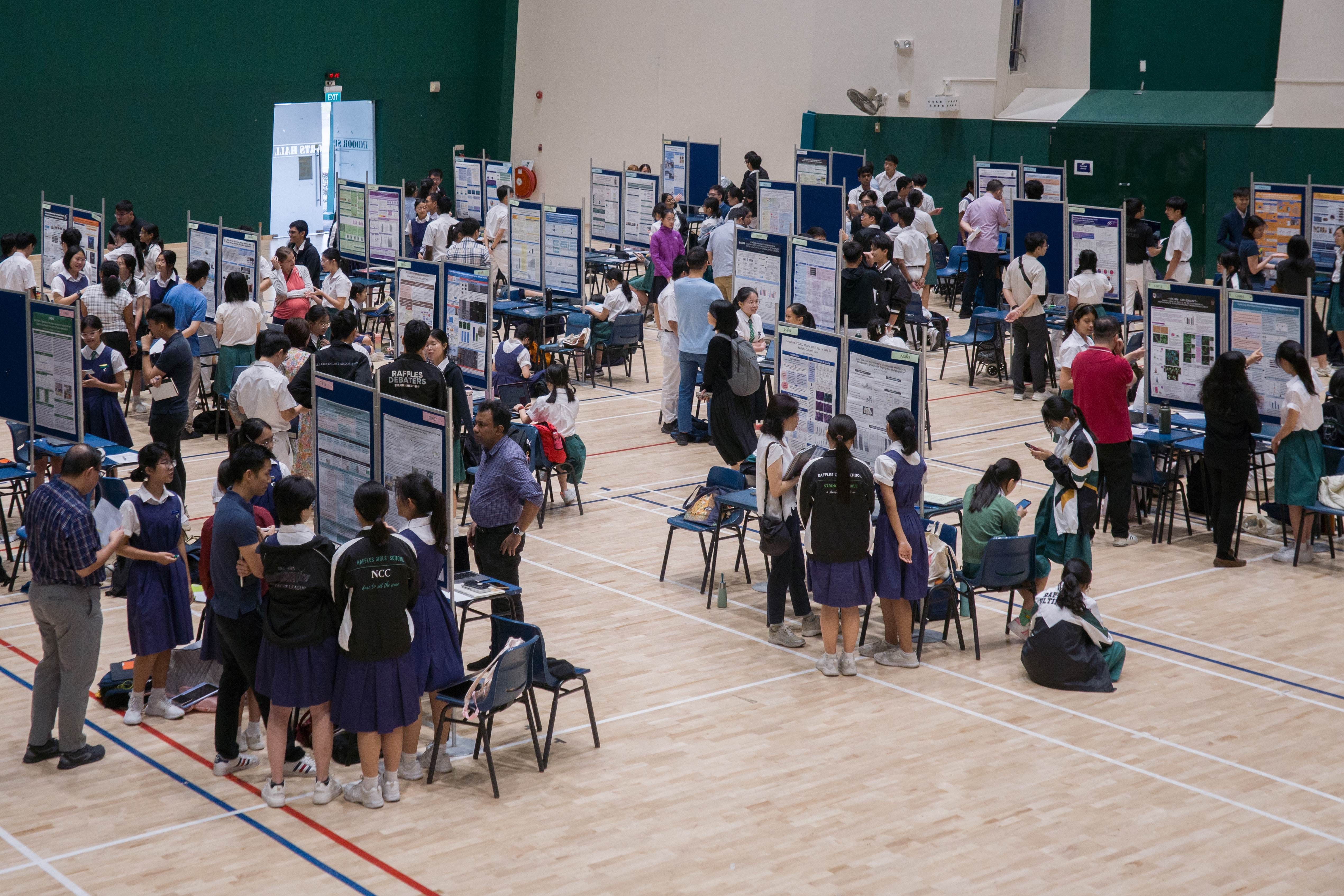 Students in school uniforms browse research poster displays in a large gymnasium during a science fair.
